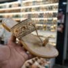 Close-up of a beige Kolhapuri sandal with intricate floral embroidery, beading and a strong toe loop, held in hand against the background of a jain chappal store display.