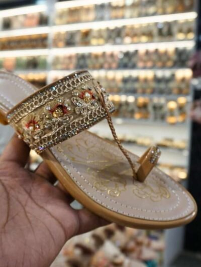 Close-up of a beige Kolhapuri sandal with intricate floral embroidery, beading and a strong toe loop, held in hand against the background of a jain chappal store display.
