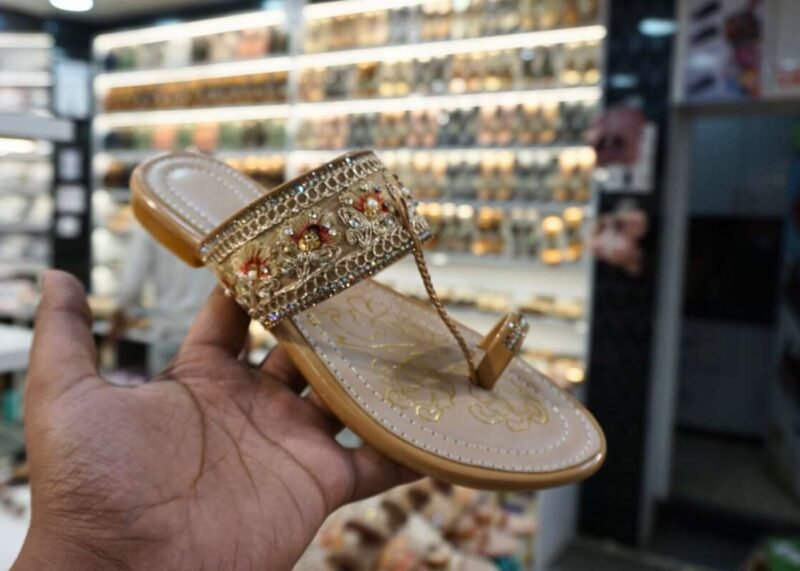 Close-up of a beige Kolhapuri sandal with intricate floral embroidery, beading and a strong toe loop, held in hand against the background of a jain chappal store display.