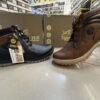 Two stylish Tiger Hill leather boots, one in black with brown accents and one in rustic brown, featuring lace-up and zipper designs, displayed on a tile floor at jain chappal store shahdara with branded packaging in the background.
