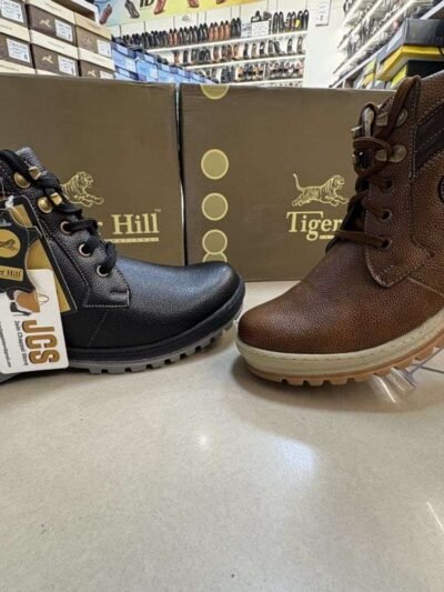 Two stylish Tiger Hill leather boots, one in black with brown accents and one in rustic brown, featuring lace-up and zipper designs, displayed on a tile floor at jain chappal store shahdara with branded packaging in the background.