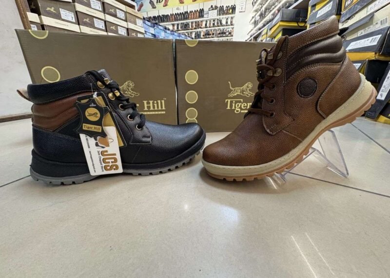 Two stylish Tiger Hill leather boots, one in black with brown accents and one in rustic brown, featuring lace-up and zipper designs, displayed on a tile floor at jain chappal store shahdara with branded packaging in the background.