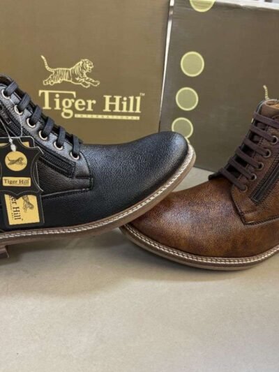 Two stylish Tiger Hill leather boots, one in black with brown accents and one in rustic brown, featuring lace-up and zipper designs, displayed on a tile floor at jain chappal store shahdara with branded packaging in the background.