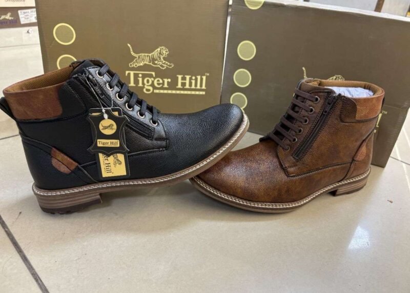 Two stylish Tiger Hill leather boots, one in black with brown accents and one in rustic brown, featuring lace-up and zipper designs, displayed on a tile floor at jain chappal store shahdara with branded packaging in the background.
