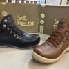 Two stylish Tiger Hill leather boots, one in black with brown accents and one in rustic brown, featuring lace-up and zipper designs, displayed on a tile floor at jain chappal store shahdara with branded packaging in the background.