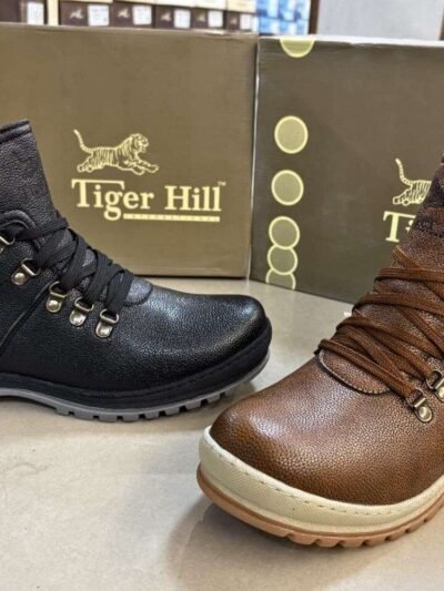 Two stylish Tiger Hill leather boots, one in black with brown accents and one in rustic brown, featuring lace-up and zipper designs, displayed on a tile floor at jain chappal store shahdara with branded packaging in the background.