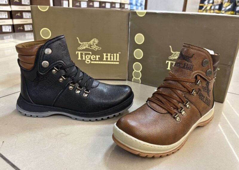 Two stylish Tiger Hill leather boots, one in black with brown accents and one in rustic brown, featuring lace-up and zipper designs, displayed on a tile floor at jain chappal store shahdara with branded packaging in the background.