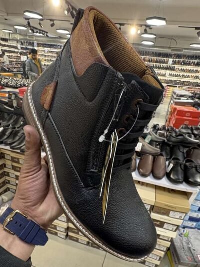Two stylish Tiger Hill leather boots, one in black with brown accents and one in rustic brown, featuring lace-up and zipper designs, displayed on a tile floor at jain chappal store shahdara with branded packaging in the background.