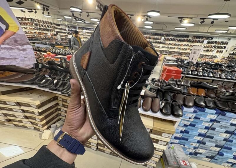 Two stylish Tiger Hill leather boots, one in black with brown accents and one in rustic brown, featuring lace-up and zipper designs, displayed on a tile floor at jain chappal store shahdara with branded packaging in the background.