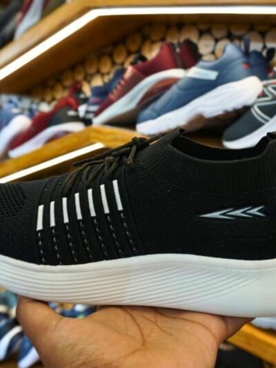 Display of sports shoes on wooden shelves in a store, with a hand holding black and white lightweight sports shoes with modern design and breathable materials, surrounded by different colored shoe options in the background.