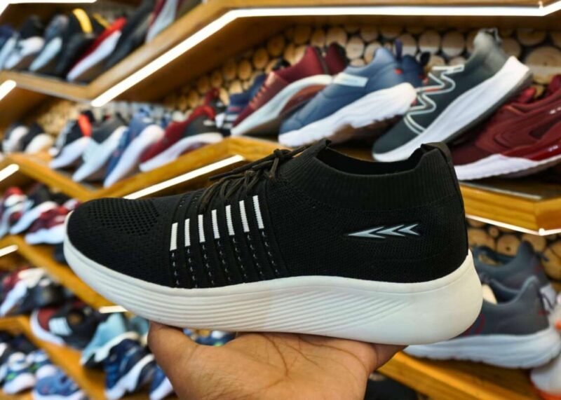Display of sports shoes on wooden shelves in a store, with a hand holding black and white lightweight sports shoes with modern design and breathable materials, surrounded by different colored shoe options in the background.