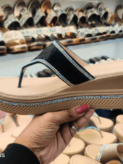 A stylish women’s platform sandal with a glossy black strap, cushioned footbed, and rhinestone-embellished sole, displayed in a footwear store.